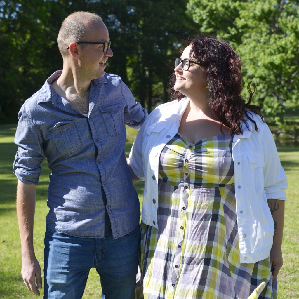 Maranda, a plus size woman wearing a yellow plaid dress and white jean jacket,  stands outdoors with her husband smiling at each other.