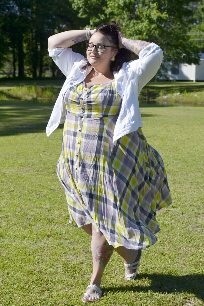 Plus size woman standing in the sun with hands in hair wearing a plaid Torrid dress with white denim jacket for Outfit of the Day.