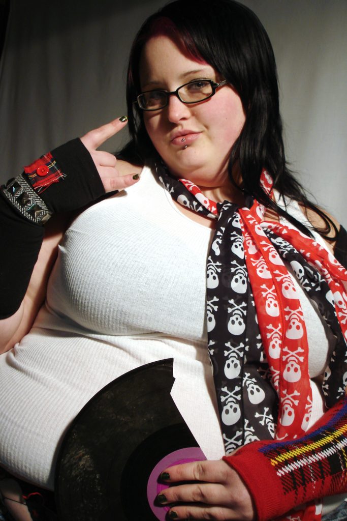 Chubby punk girl with long black hair wearing skull print scarves and punk accessories in dramatic lighting.