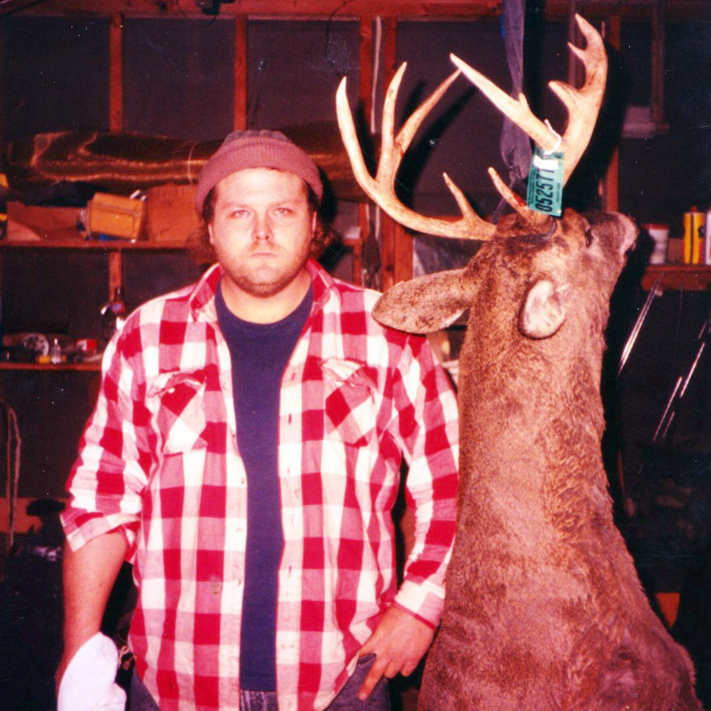 Man wearing flannel shirt stands next to hanging trophy deer.
