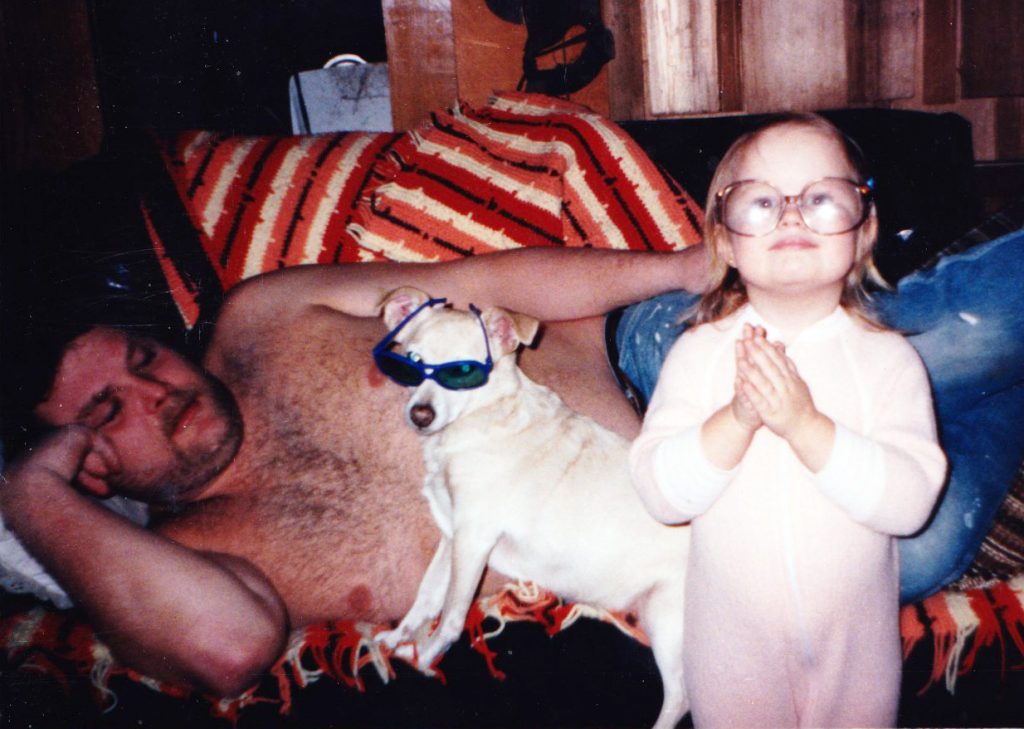 Young Maranda in pajamas wearing oversized glasses. Behind her, a small white dog wearing sunglasses and her dad laying on a couch.