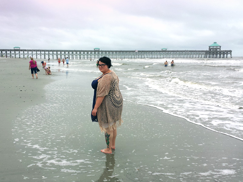 Maranda a plus size woman, standing near the water at Folly Beach in South Carolina. Pier 101 spans the background horizon.