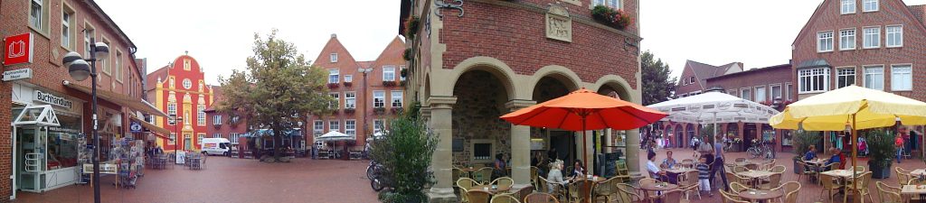 Panoramic view of historic market plaza in Meppen, Germany, featuring brick architecture and umbrellas over outdoor dining tables.
