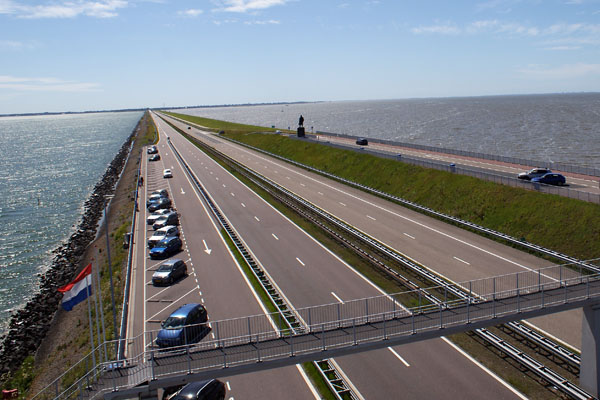 View into the distance of the Afsluitdijk in the Netherlands showing roadways in both directions between sparkling water and open blue sky.