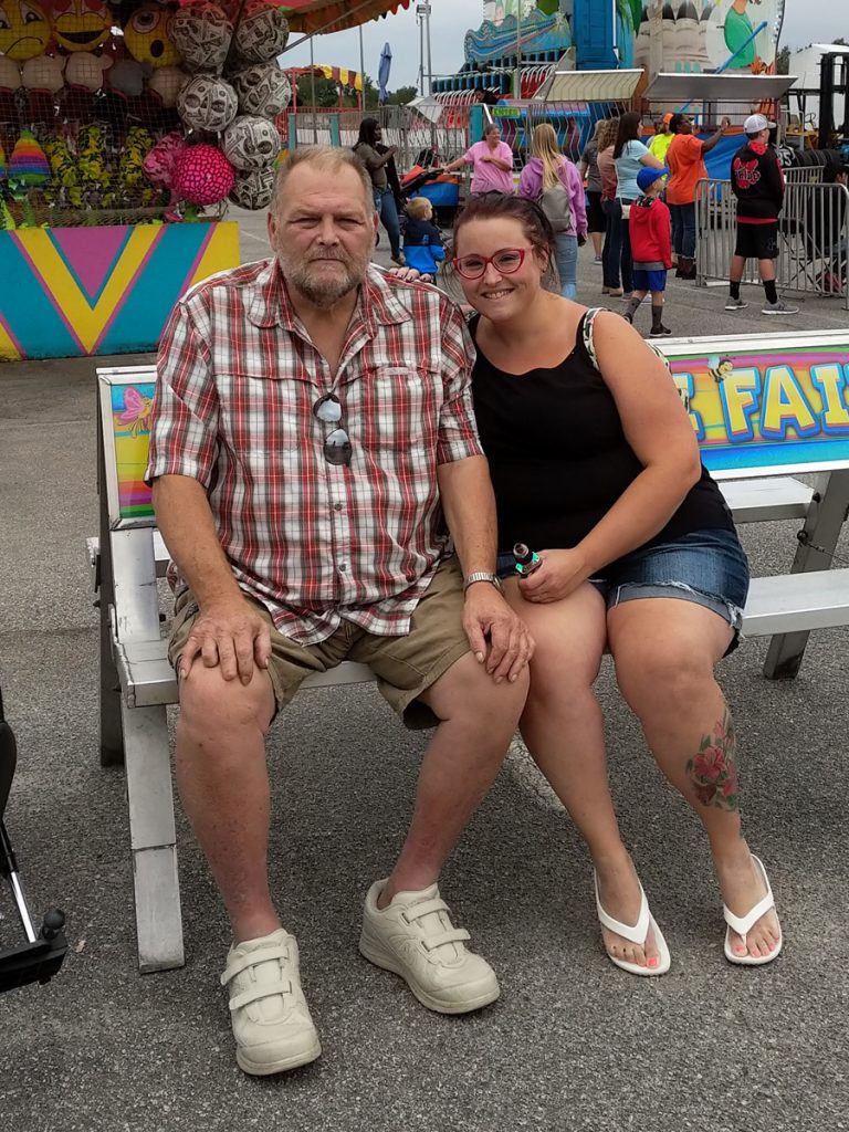 Maranda sits with her dad outdoors at a carnival.