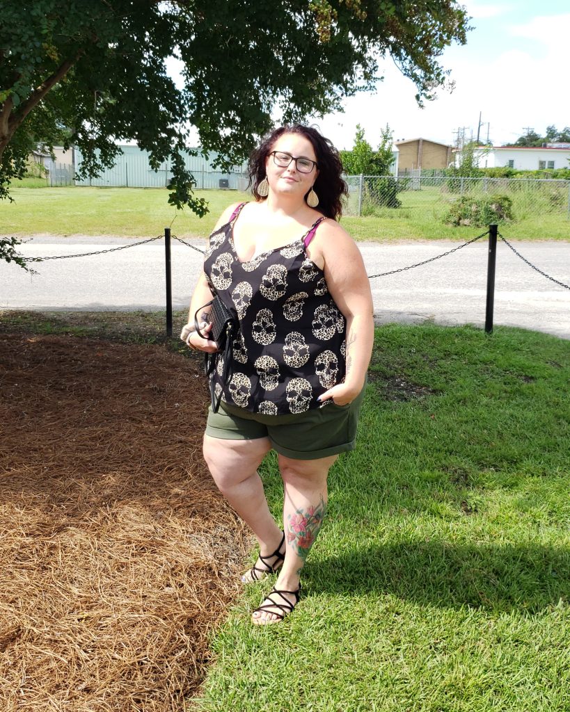 Maranda, a plus size woman, stands outdoors wearing a black and leopard skull print tank top, olive green shorts, and black strappy sandals.