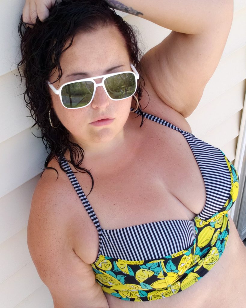 Maranda, a plus size woman, stands outdoors against a wall wearing aviator sunglasses and a lemon print bikini top from Torrid.