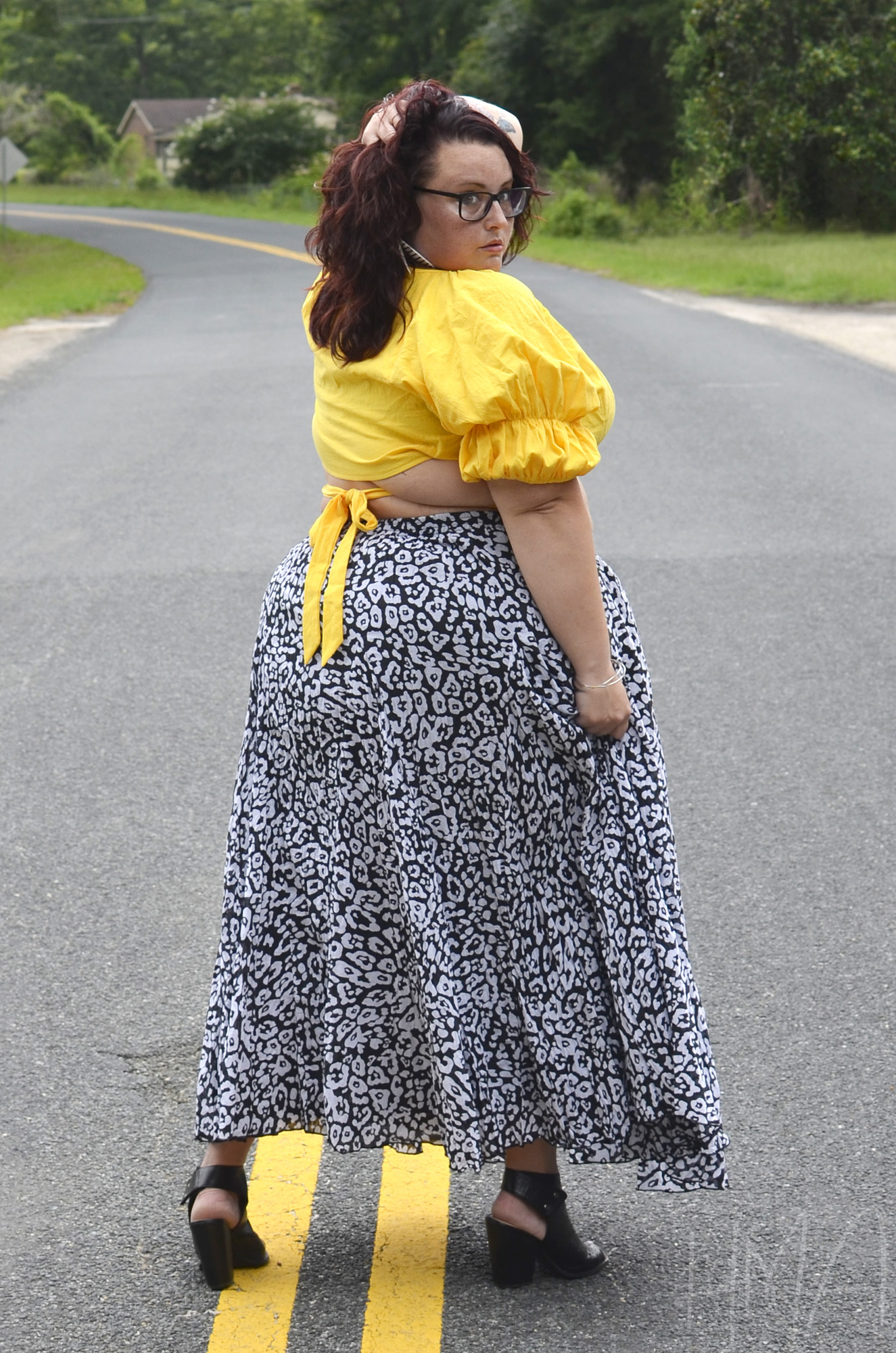 Maranda, a plus size woman, models a leopard print maxi skirt and yellow crop top from the back, looking over her shoulder at the camera.