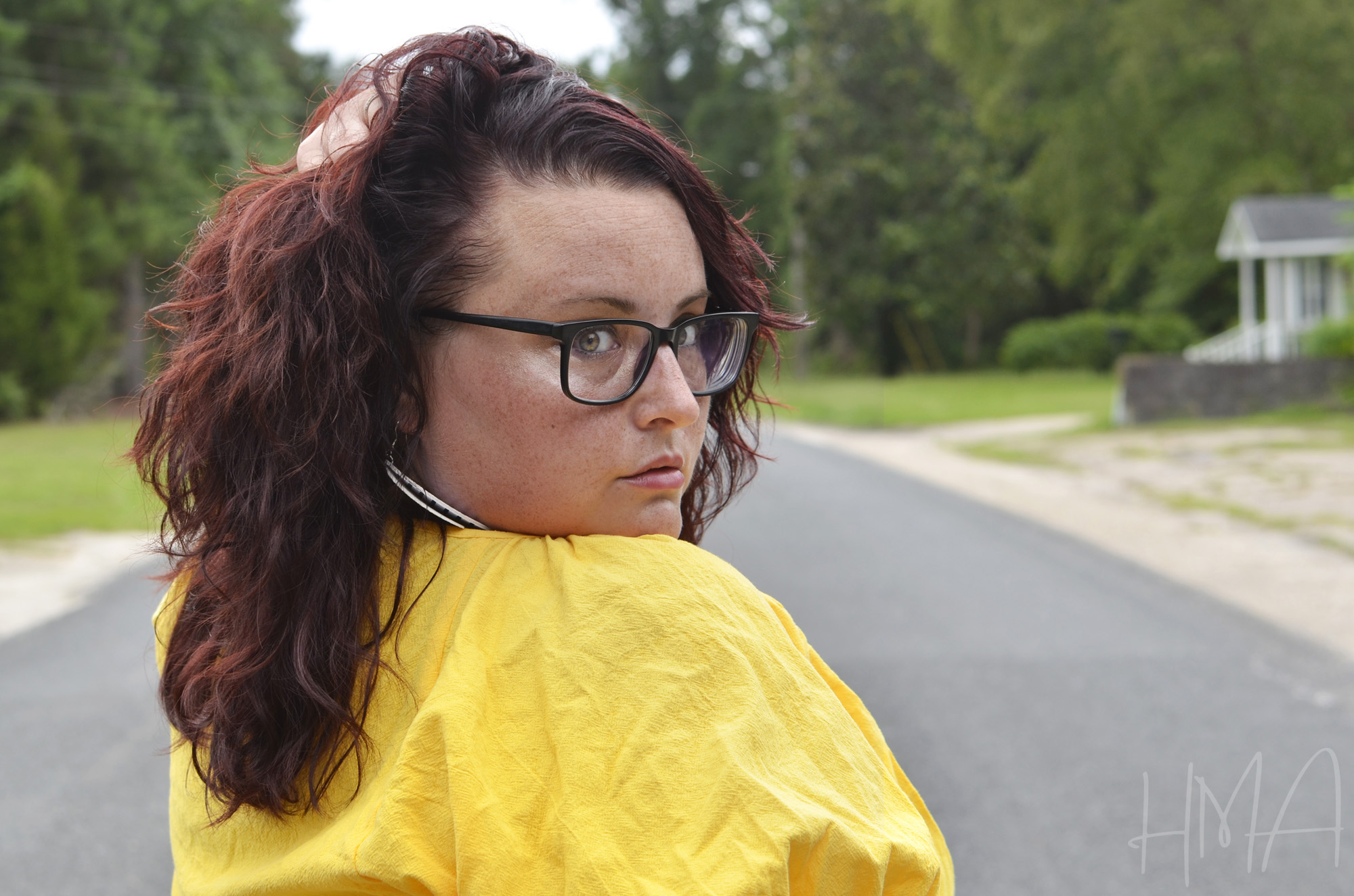 Maranda faces away from the camera and looks back over her shoulder, wearing nerdy black glasses and a yellow top.