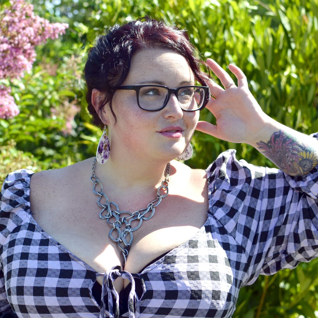 Maranda, a plus size woman, models a black and purple gingham muse dress from Wild Fable and chainmail necklace by Cookie Lee.
