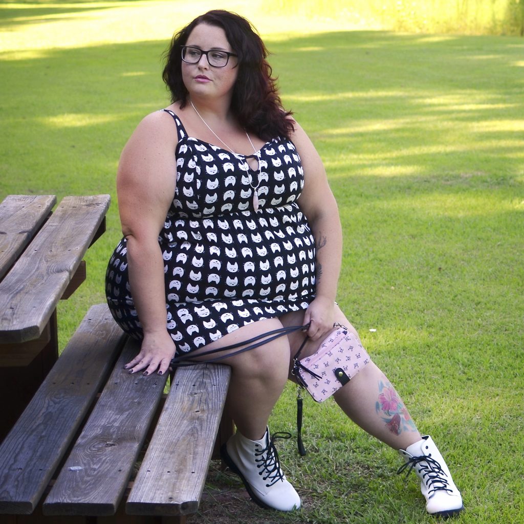 Maranda, a plus size woman, sits on a wooden bench wearing a black and white cat face print sun dress and white combat boots.