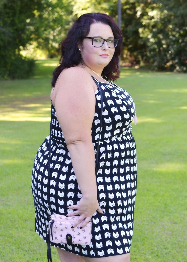 Maranda, a plus size woman, stands outdoors on a sunny day, looking at the camera from a side profile that shows her black and white plus size cat print dress down to the knees.