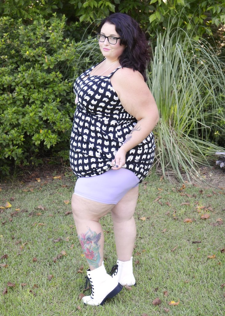 Maranda, a plus size woman, stands outdoors in profile to the camera, pulling her black and white dress up at the hip to reveal pastel purple Yitty shapewear shorts underneath.