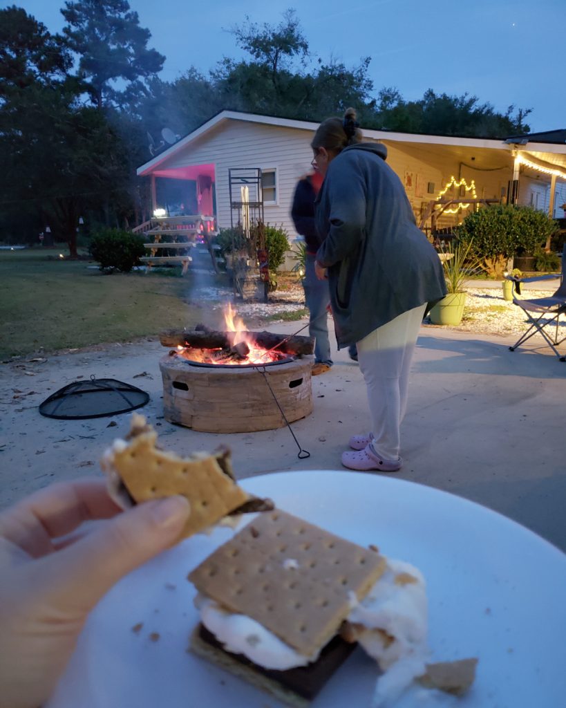 S'mores on a plate in the foreground, with Mom standing in the background tending a campfire in a round fire pit.