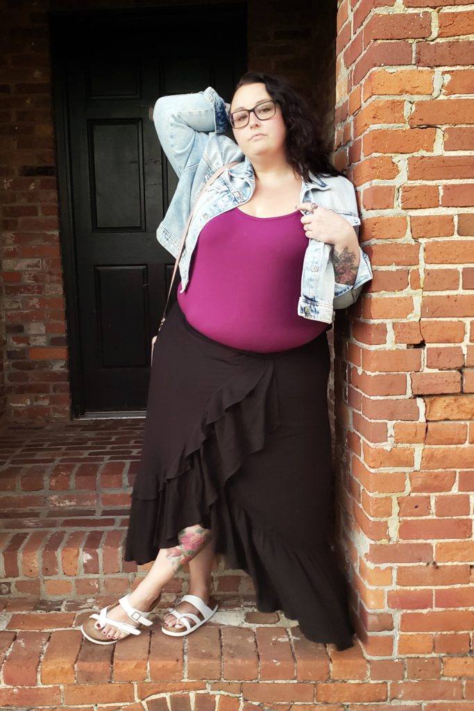 Maranda, a plus size woman, stands outdoors in an old downtown brick doorway wearing a black ruffle hi-lo maxi skirt and plum top from Torrid, topped with an acid washed denim jacket.