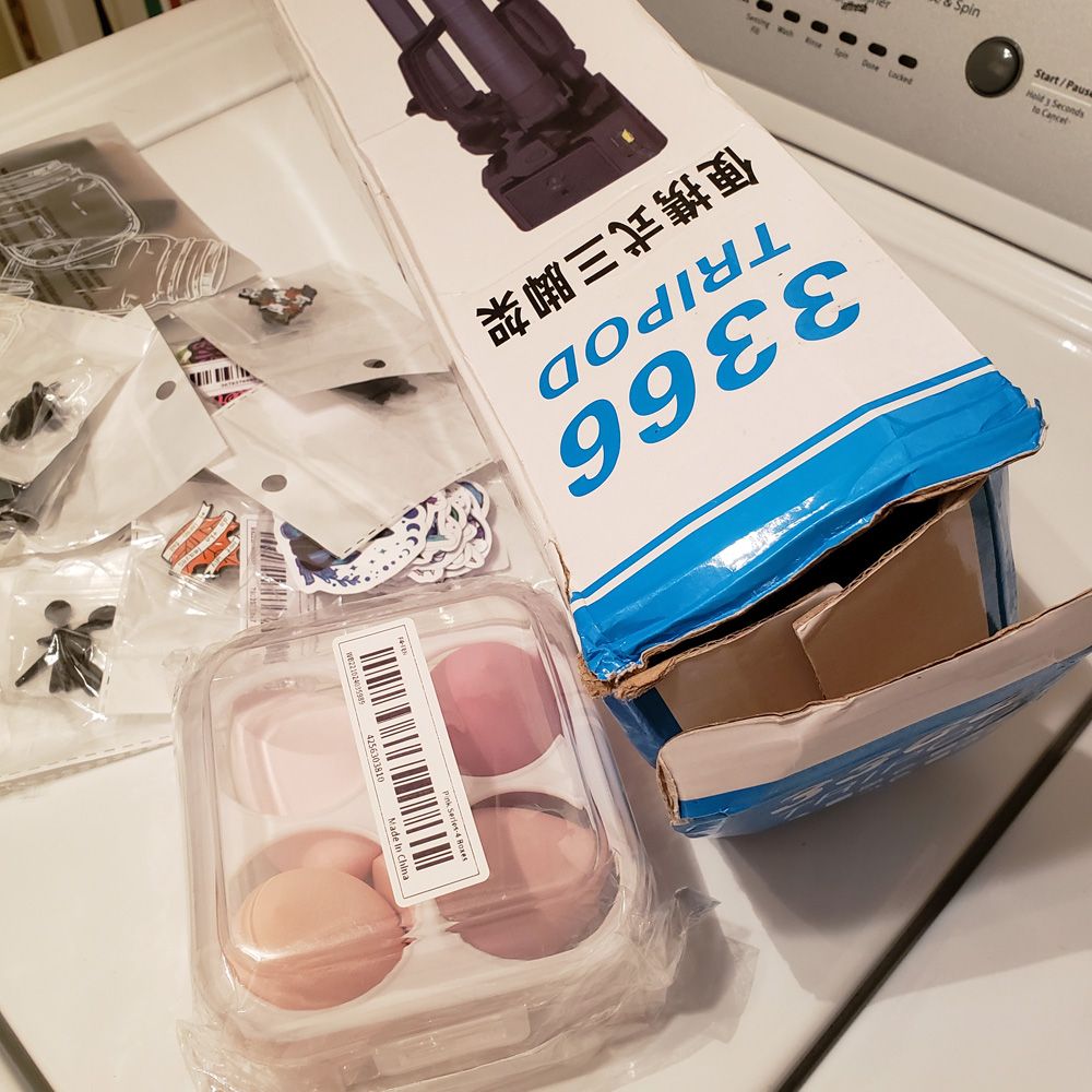 A collection of enamel pins in packages, a set of makeup sponges, and a cardboard box for a tripod are grouped together as received from Temu.