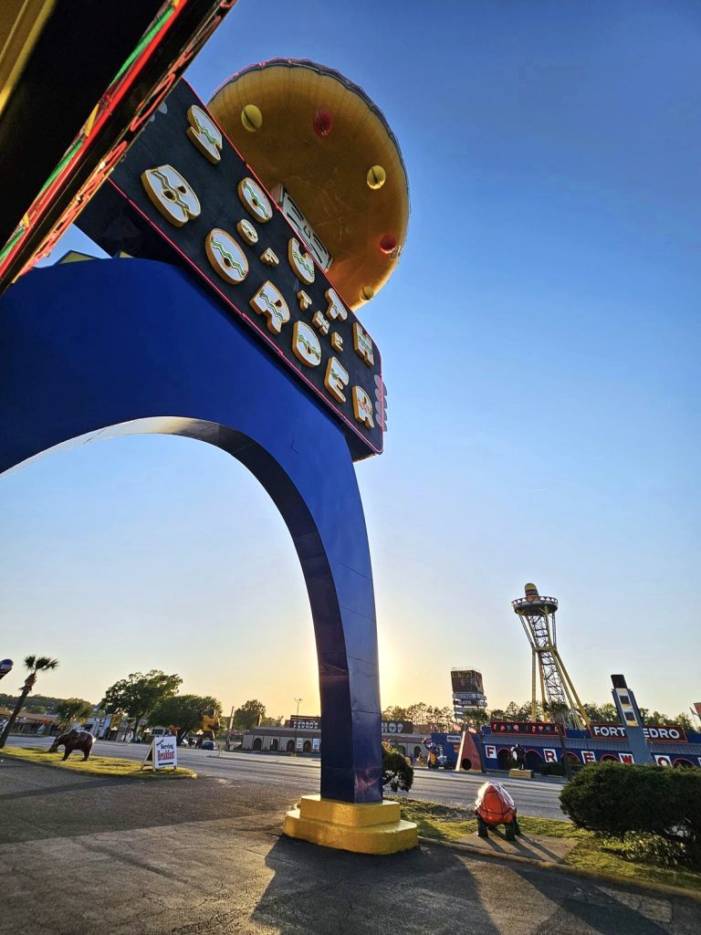 Recent-day photo taken underneath the Pedro archway, looking up at the South of the Border logo signage and neon lights. The sombrero tower is silhouetted by the sunset in the background.