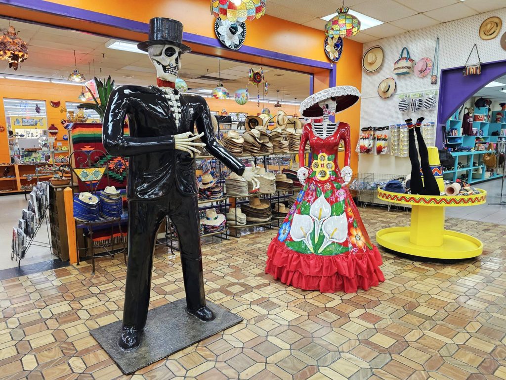 Calavera skeleton metal larger-than-life statues greet visitors near the front of the Mexico Shop East store, surrounded by a large hat display and gifts for sale.