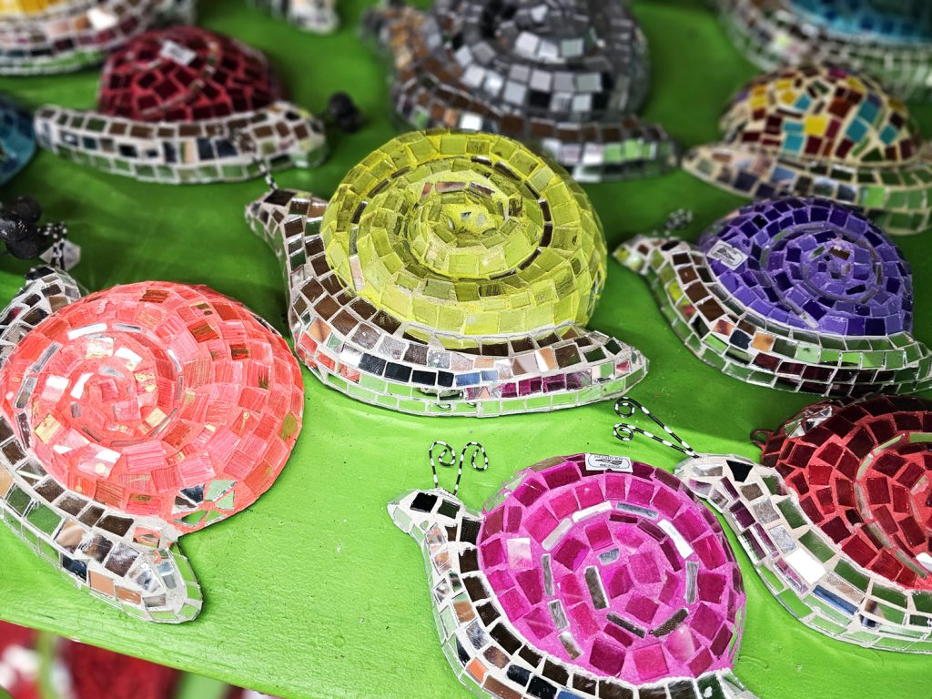 Mexican-made ceramic garden stones shaped like snails are adorned with vibrant paint colors and mirrored glass tile mosaics.