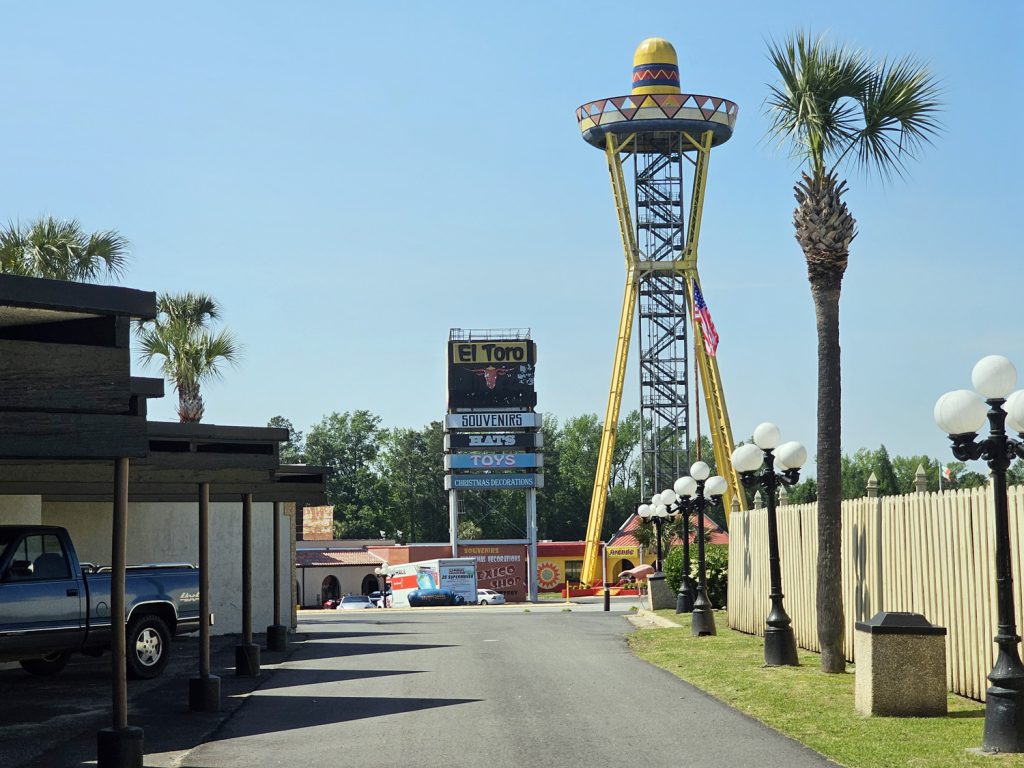 View from behind a South of the Border Motor Inn room carport, including the sombrero tower and parking lot, under a clear blue sky.