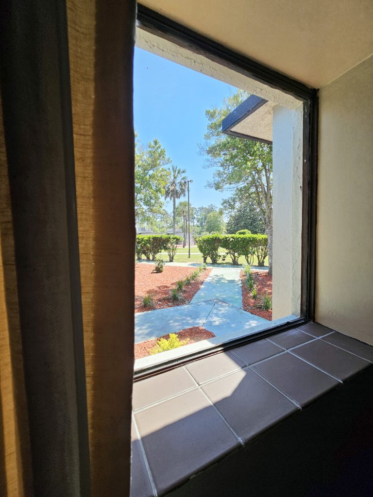 Interior guestroom view at South of the Border Motor Inn, looking outside to a blue sky, sunny day, and walking paths neatly trimmed.