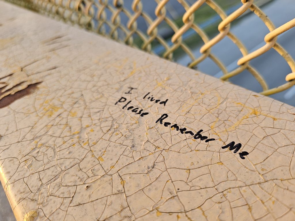 Close-up, "I lived; please remember me" written in black permanent marker on the chippy yellow paint of the South of the Border walkway.