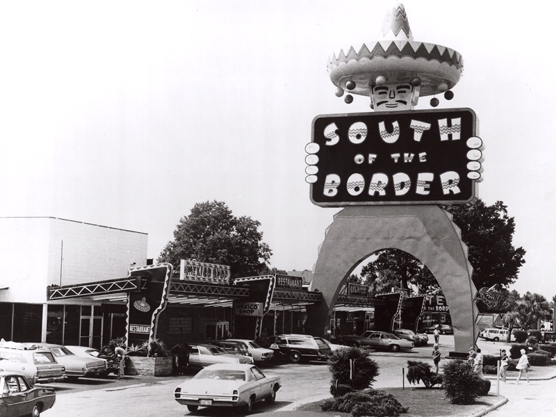 Black and white photo, circa 1960's, of automobiles and tourists within the South of the Border roadside attraction, towered by the iconic Pedro archway displaying the establishment's logo.